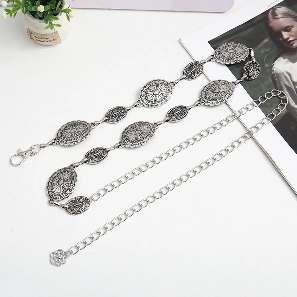 Cadena de cintura ovalada de plata delicada con eslabones decorativos, mostrada sobre un fondo blanco junto a un libro.
