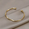 A minimalist gold-tone twisted cuff bangle resting on a neutral gray fabric.