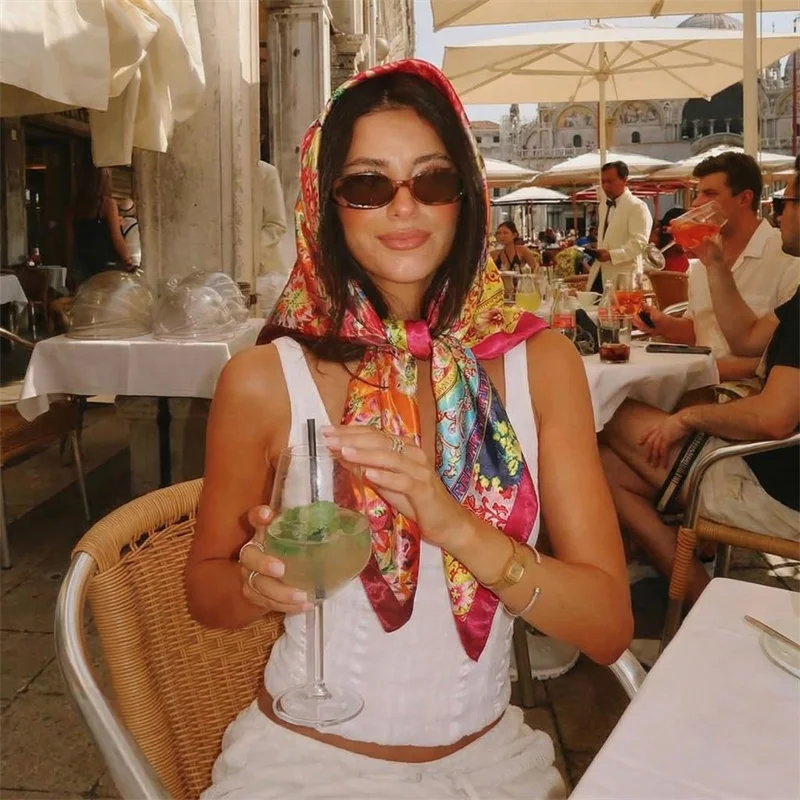 Eine Frau mit buntem Seiden-Satin-Hijab und Sonnenbrille sitzt an einem Außencafé-Tisch.