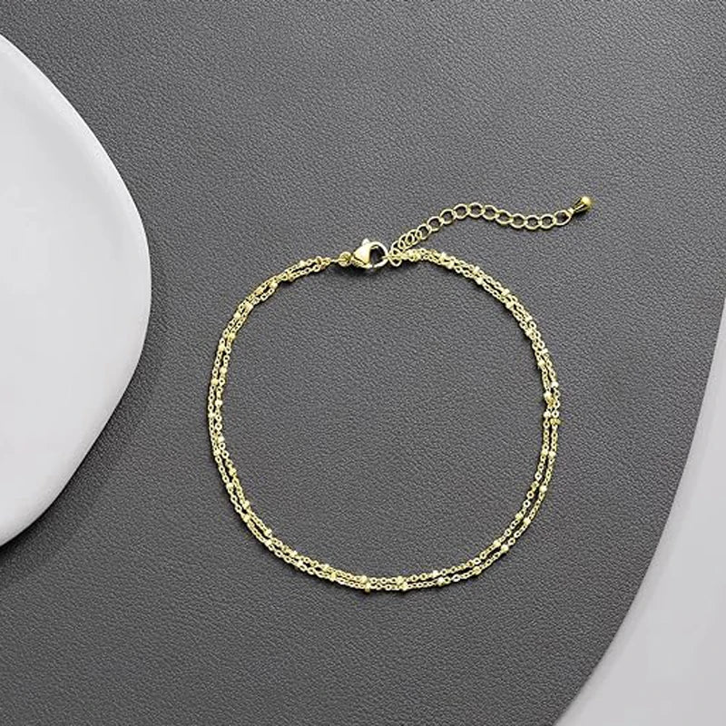 A delicate layered gold and silver plated anklet is displayed on a gray surface.