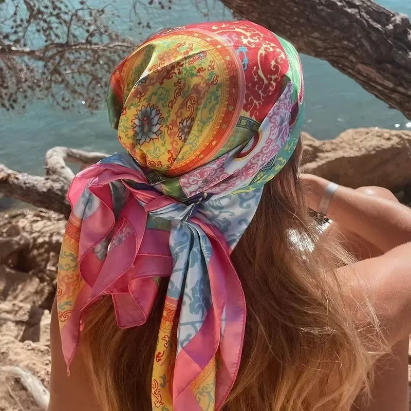 A woman wearing a colorful silk square scarf tied around her head, outdoors near the sea.