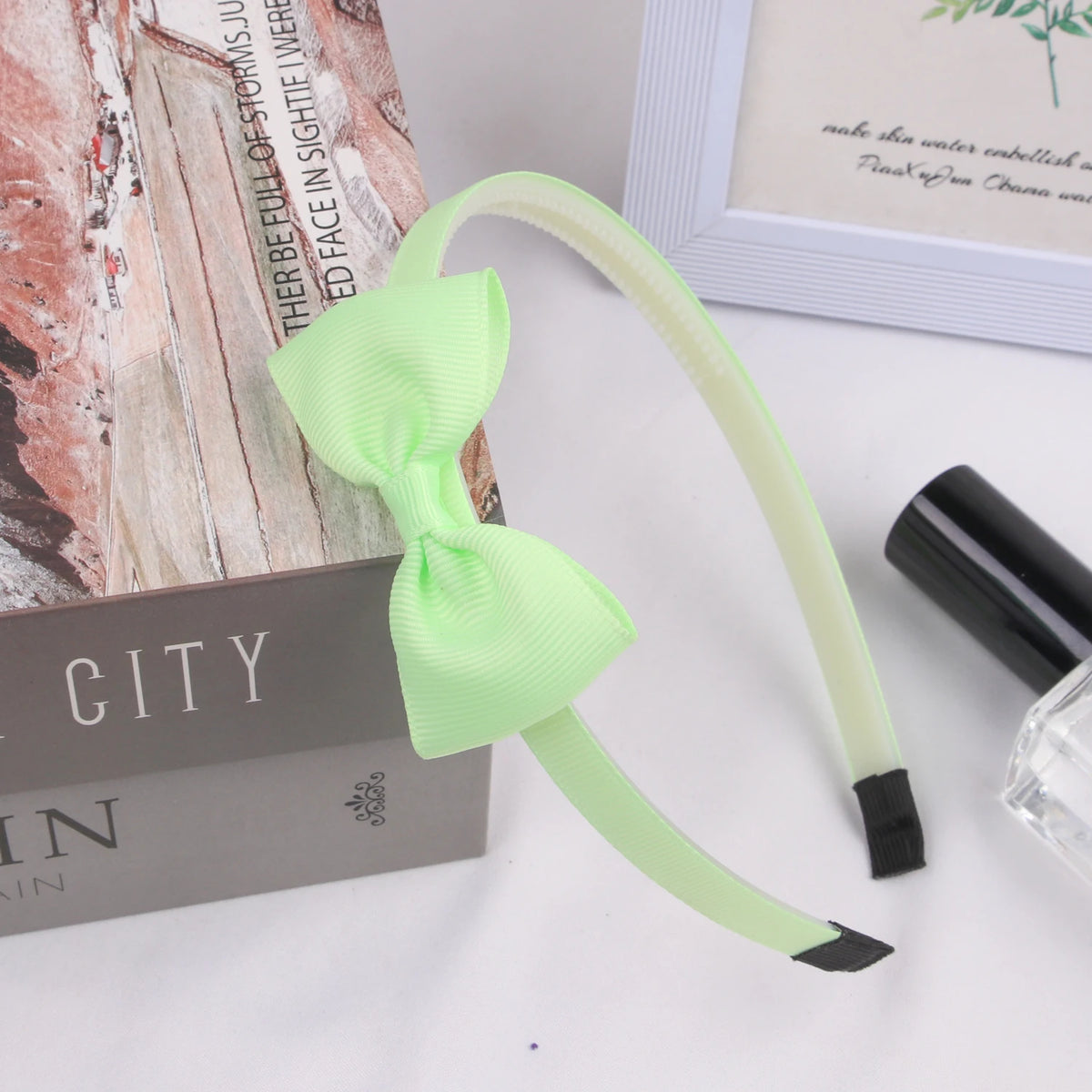 A light green grosgrain ribbon hair bow is displayed next to a cosmetic bottle and a book.