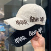 A hand holds a white baseball cap and a black baseball cap both embroidered with the words NEVER GIVE UP