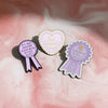 A set of three enamel pins featuring a heart shape, a ribbon, and a medal design on a pink background.