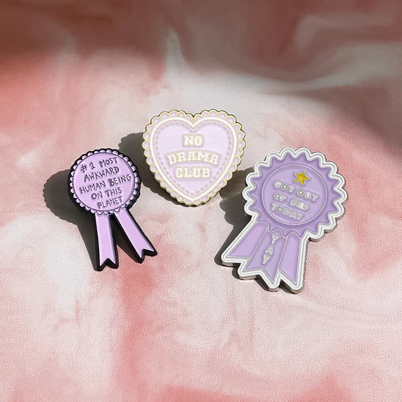A set of three enamel pins featuring a heart shape, a ribbon, and a medal design on a pink background.