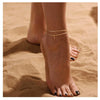 A close-up of a woman's ankle showing layered letter anklets made of gold-toned stainless steel charms against a sandy backgr