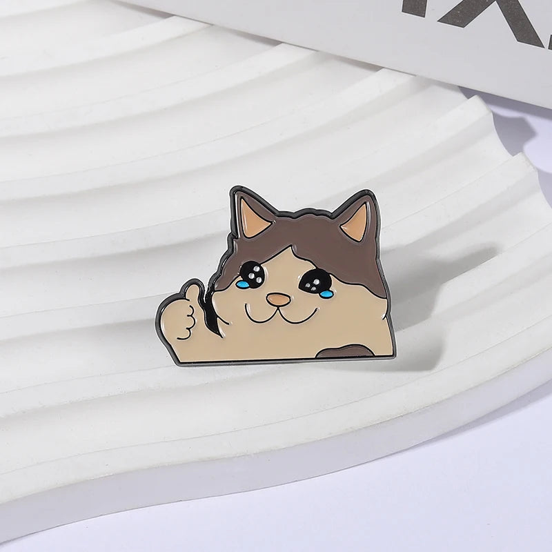 A cute cat face enamel pin with blue eyes and a smiling expression resting on a white surface.