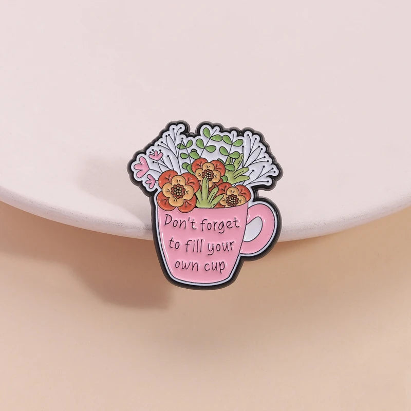 A pink enamel brooch shaped like a teacup filled with flowers and the text Don't forget to fill your own cup