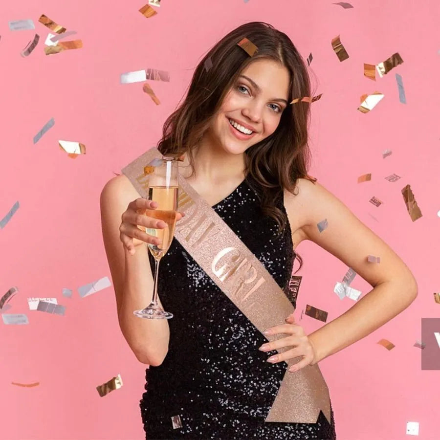 A smiling young woman wearing a sparkly black dress holds an elegant gold sash and a glass of amber liquid against a pink bac