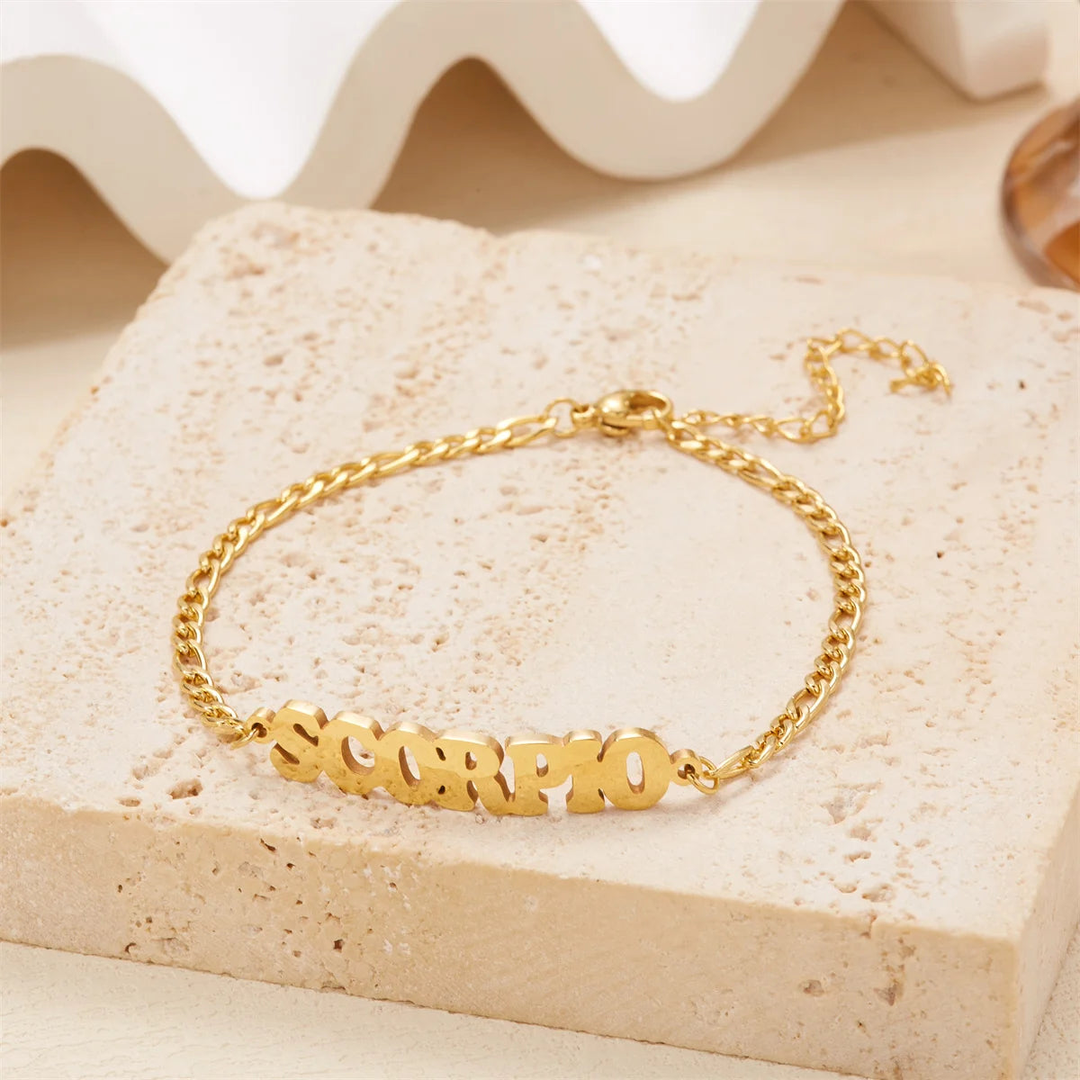 A gold stainless steel zodiac constellation charm bracelet with the word SCORPIO resting on a light beige stone.