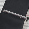 A silver-toned business tie clip with a bar clasp is shown resting on a dark, textured surface.