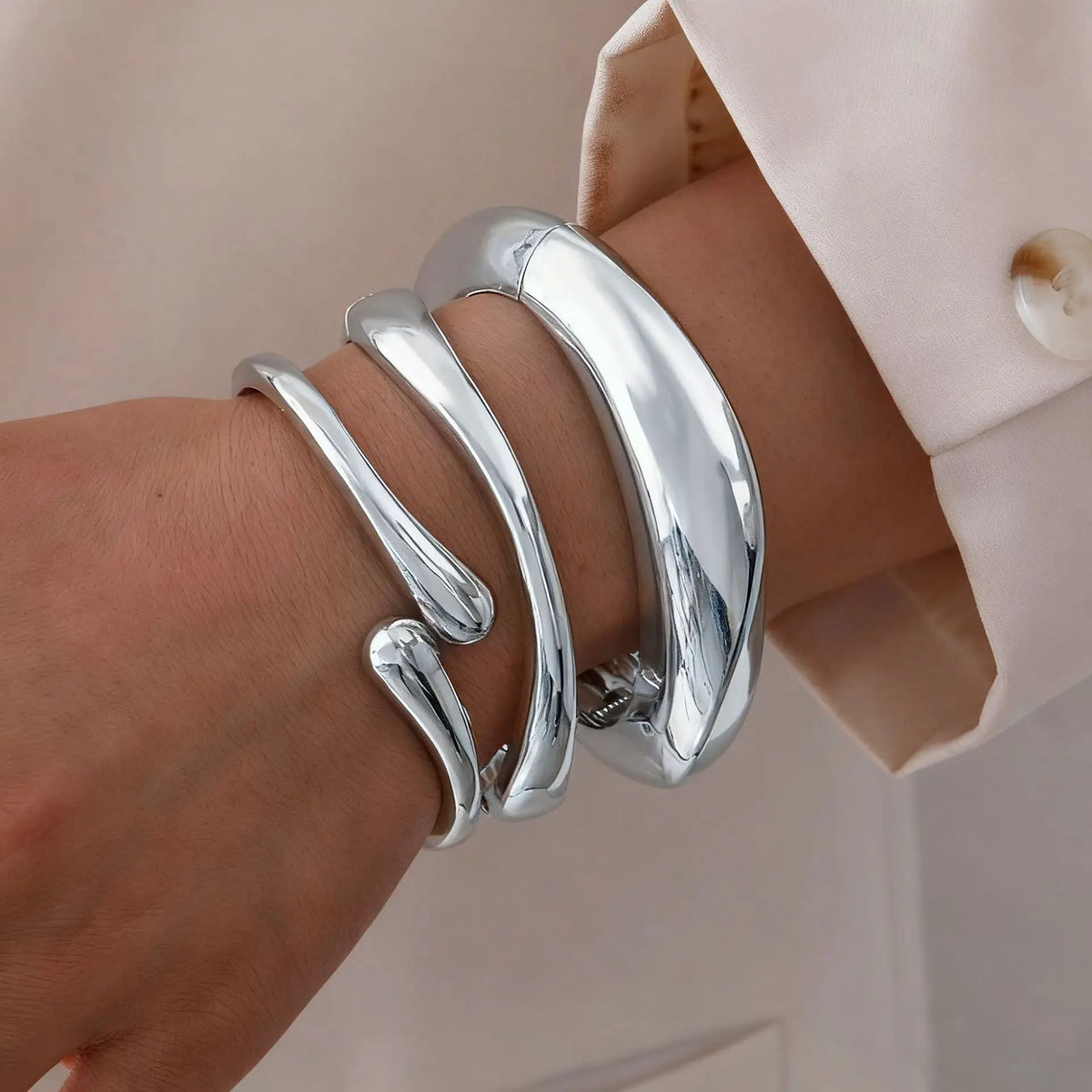 A close-up of a hand wearing a stack of silver chunky cuff bangle bracelets on a wrist, next to a light pink blazer sleeve.