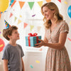 A smiling woman gives a boy a gift box in a bright, festive indoor setting.