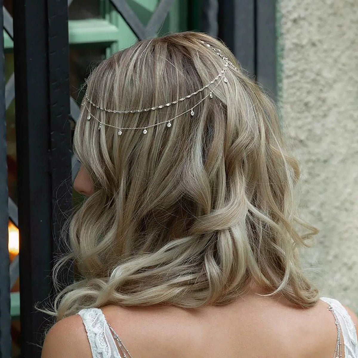 A woman with light blonde hair shows the back of her head adorned with a delicate crystal chain bridal hair accessory.