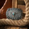Boucle de ceinture en métal argenté avec tête de taureau détaillée et incrustation turquoise, posée sur un sac de jute.