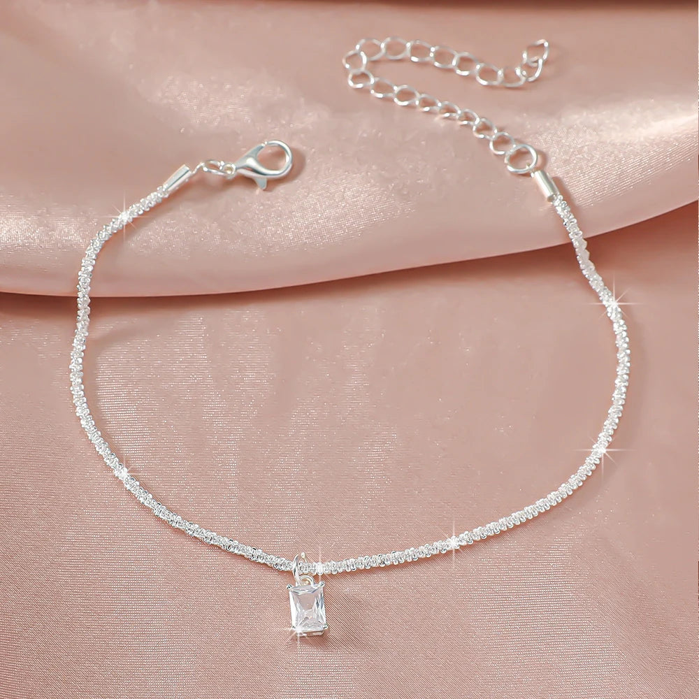 A delicate silver-plated chain anklet with a sparkling crystal charm displayed on a pink fabric background.