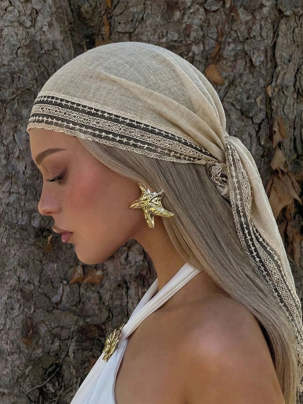 A model wearing a beige bohemian lace trim shawl scarf and gold earrings, looking to the side against a tree trunk.
