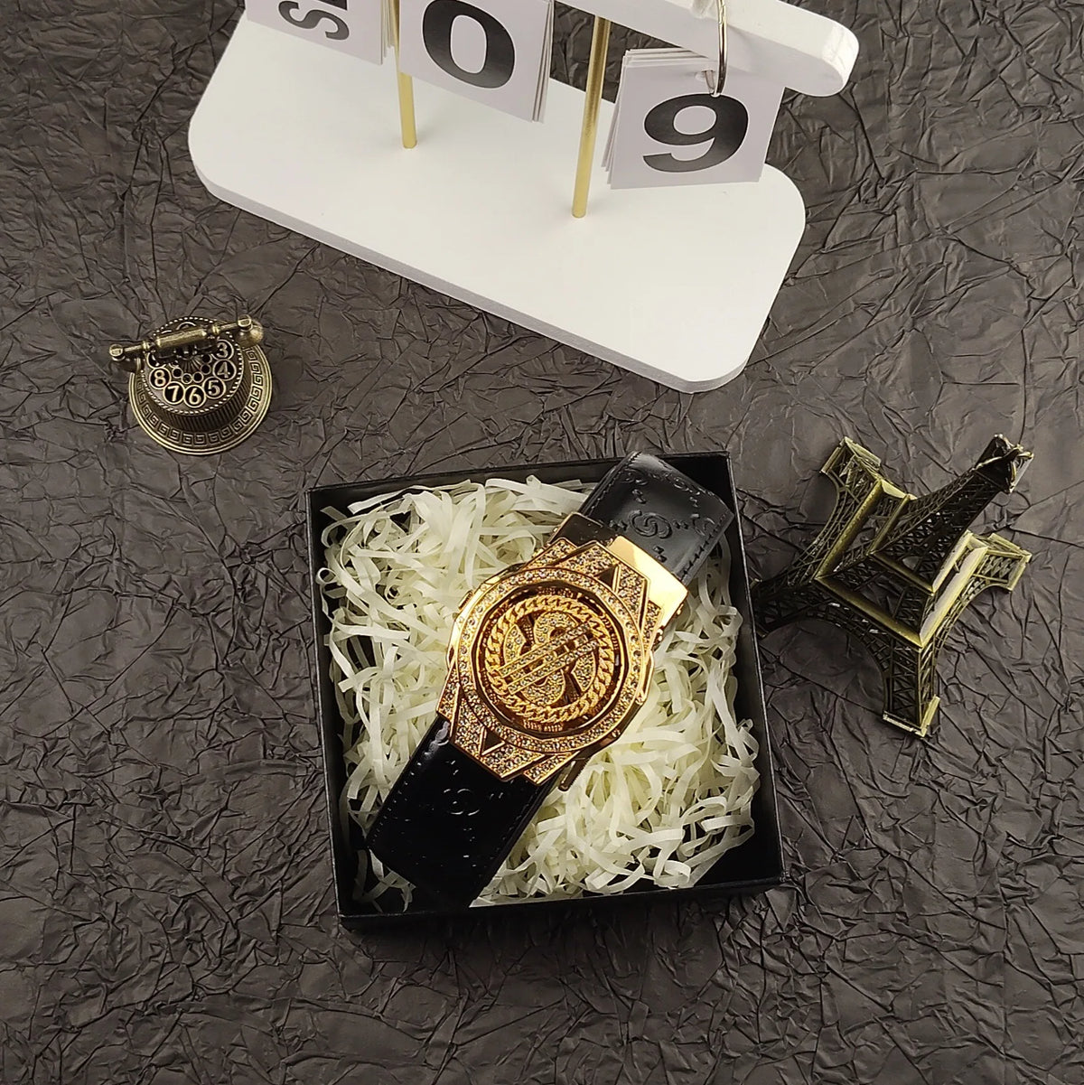 A luxury gold-toned watch is placed on a bed of white shredded material next to a black leather belt and a gold-toned buckle.