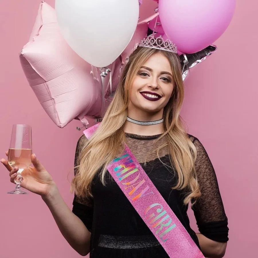 A young woman with long blonde hair smiles while holding a glass of pink beverage and surrounded by pink balloons and a glitt