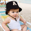 A baby wearing a black sun hat with an embroidered letter A is relaxing in a striped beach lounge chair.