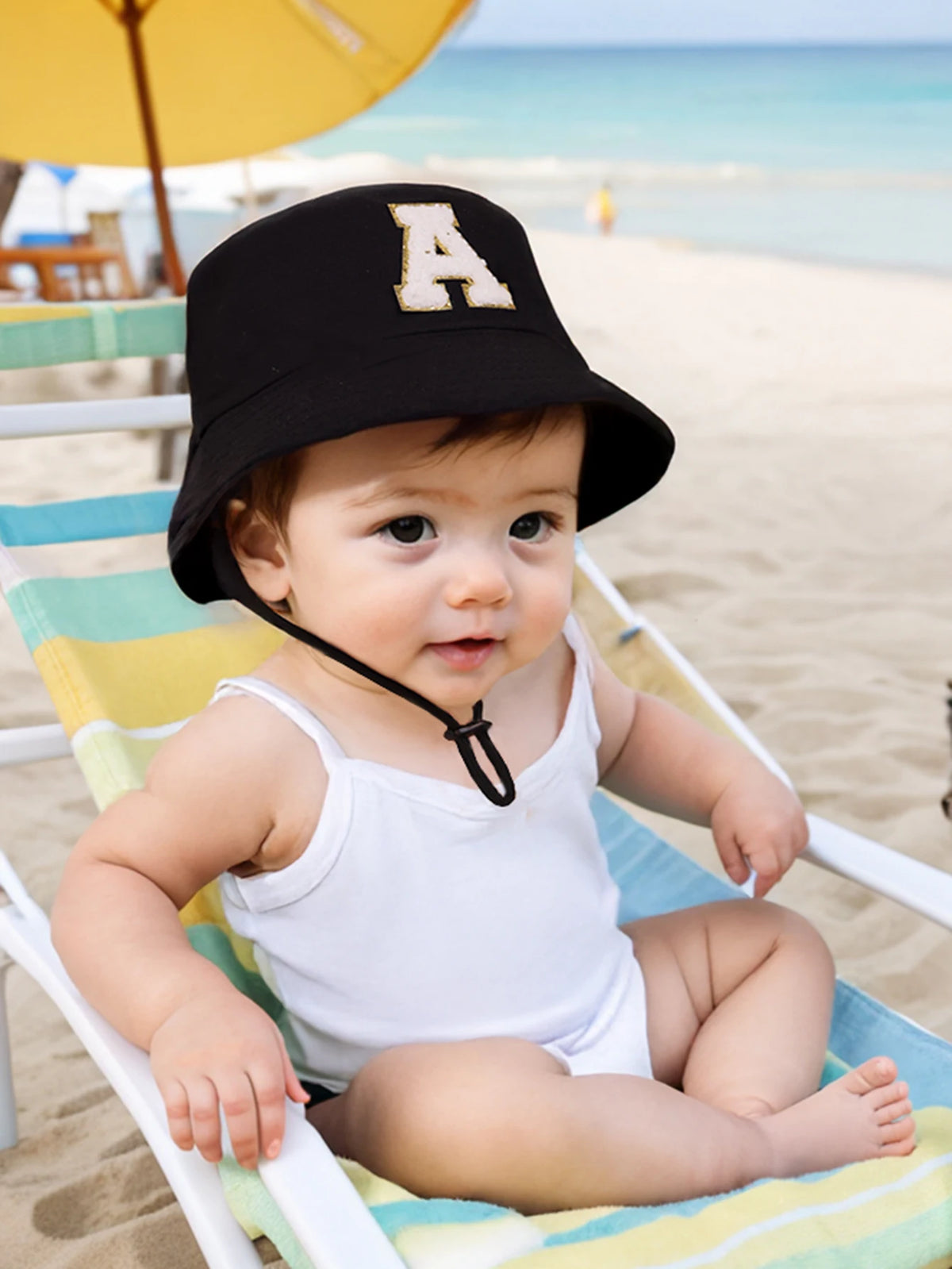 A baby wearing a black sun hat with an embroidered letter A is relaxing in a striped beach lounge chair.