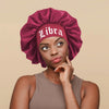 A woman wearing a deep magenta satin sleep bonnet with a wrap band that spells Libra on the front.