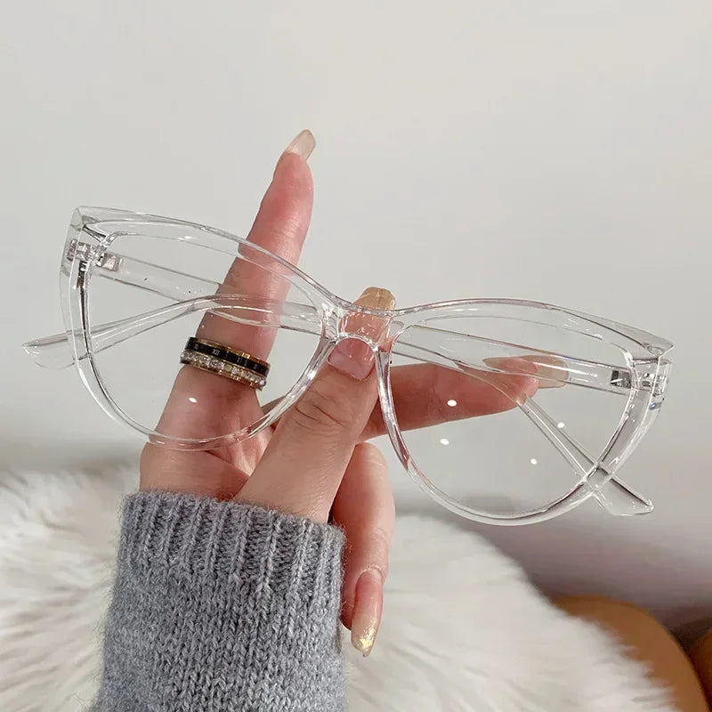 Eine Hand hält eine klare Vintage Cat-Eye-Brille mit goldenen Akzenten vor einem weißen, flauschigen Hintergrund.