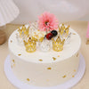 A white cake decorated with gold and silver mini crown toppers, a pink flower, and dark berries.