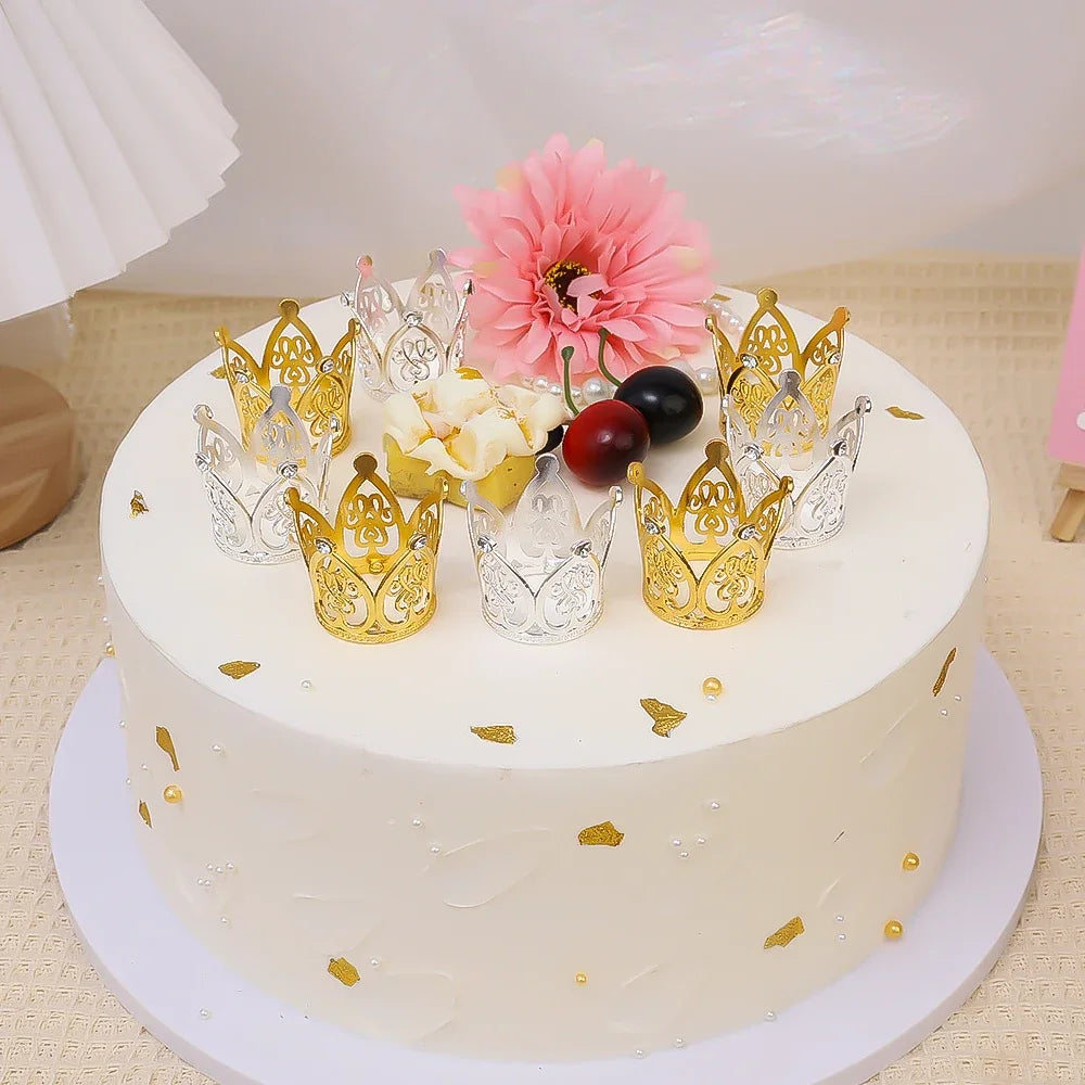 A white cake decorated with gold and silver mini crown toppers, a pink flower, and dark berries.