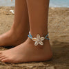 A close-up of a woman's ankle wearing a bohemian cowrie shell anklet with a white flower charm on a sandy beach.