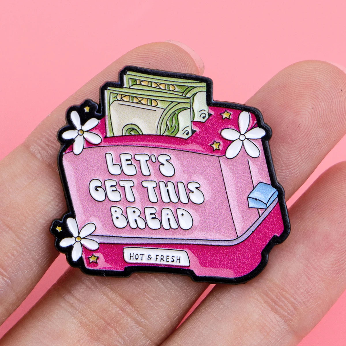 A pink enamel pin featuring a loaf of bread and the text Let's Get This Bread, held by a hand.
