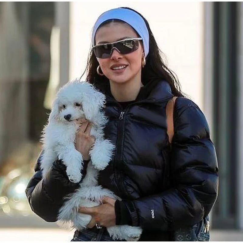A woman wearing sunglasses and a black jacket holds a white fluffy dog while wearing a blue knitted sports headband.
