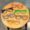 A collection of colorful square frame costume glasses in red, orange, black, green, blue, and yellow resting on a wooden tabl
