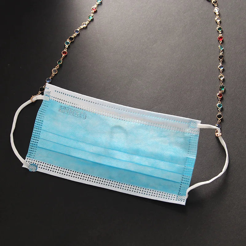 Una mascarilla desechable azul claro expuesta contra un fondo oscuro, sujeta a una cadena de cuentas de cristal ornamentada.