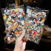 A hand holds up two clear plastic pouches filled with various enamel badge pins and decorative brooches.