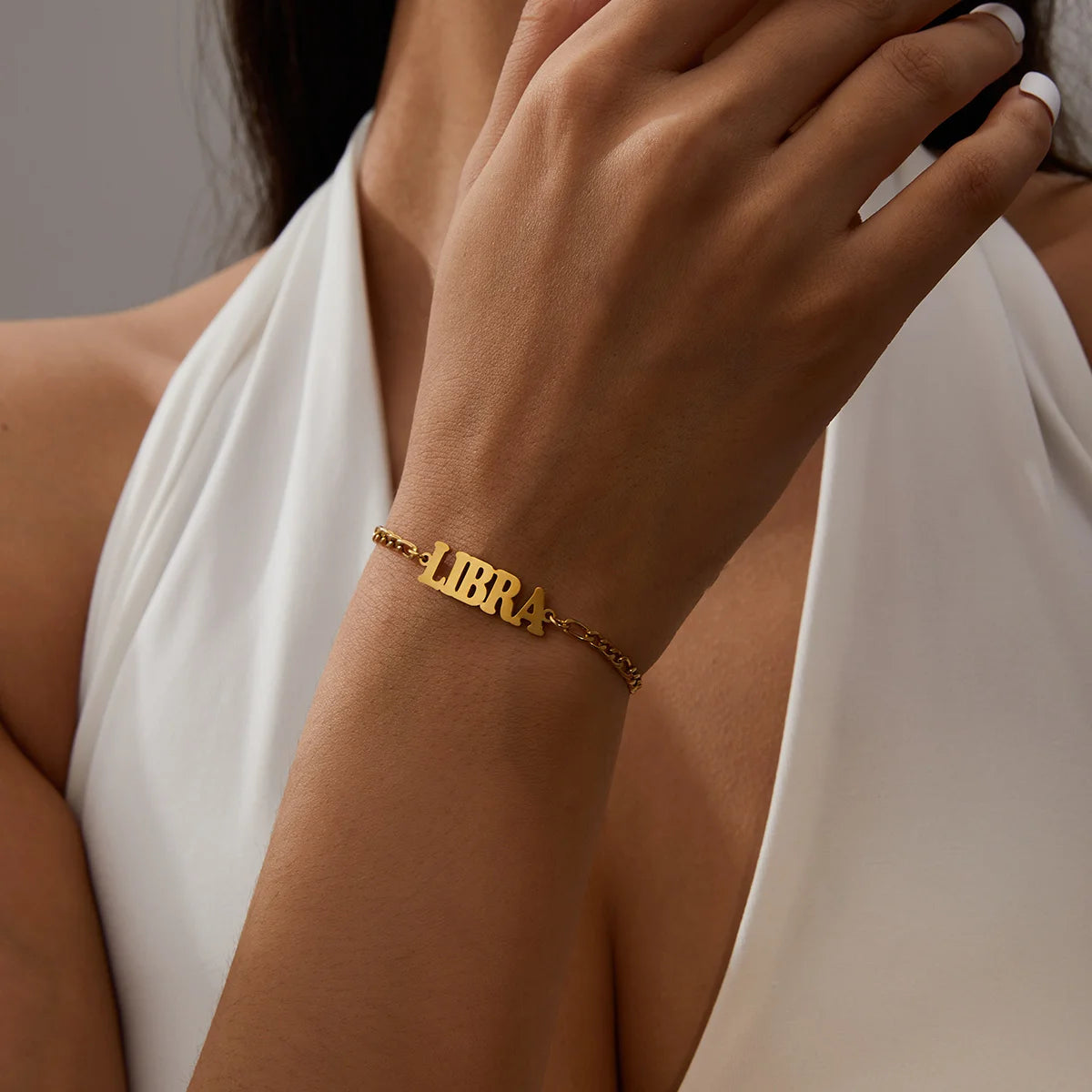 A close-up of a hand wearing a gold stainless steel bracelet engraved with the word LIBRA on a white dress.