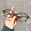 A hand holds a pair of round tortoiseshell computer glasses with green accents against a gray background.