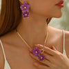 A close-up of a gold floral pendant necklace and matching earrings featuring purple flowers on a woman's neck and hand.