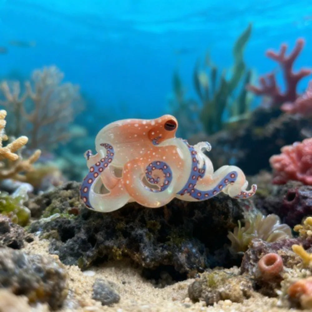 Une petite pince à cheveux en forme d'octopus orange translucide posée sur un fond de récif avec des plantes marines dans l'e