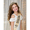 A smiling girl wearing a gold birthday tiara crown and sash against a soft, light background.