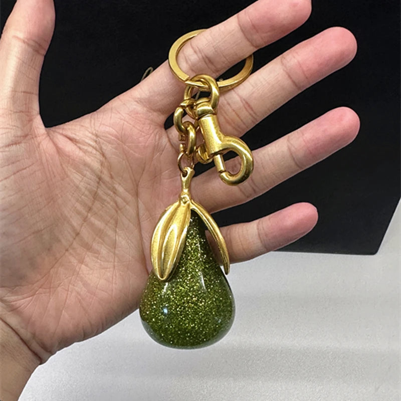 Un charm de sac en tons dorés avec un pendentif en forme de poire verte et scintillante tenu dans une main.