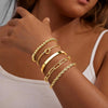 A close-up of a hand wearing several stacked 14K gold plated chunky cuff bangles on a wrist.