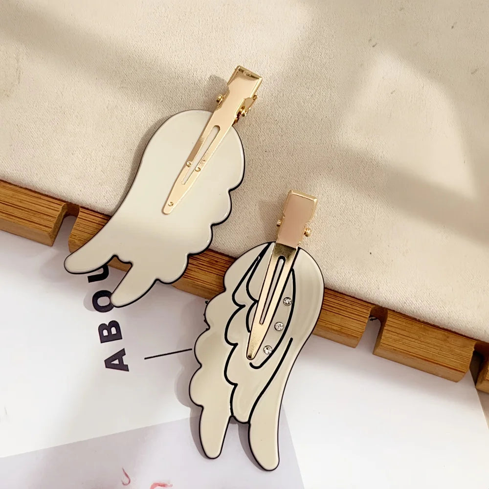 A pair of gold-toned angel wing hair clips displayed on a wooden surface.