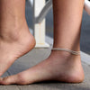 A close-up of a person's ankle wearing a delicate, silver-plated beaded anklet.