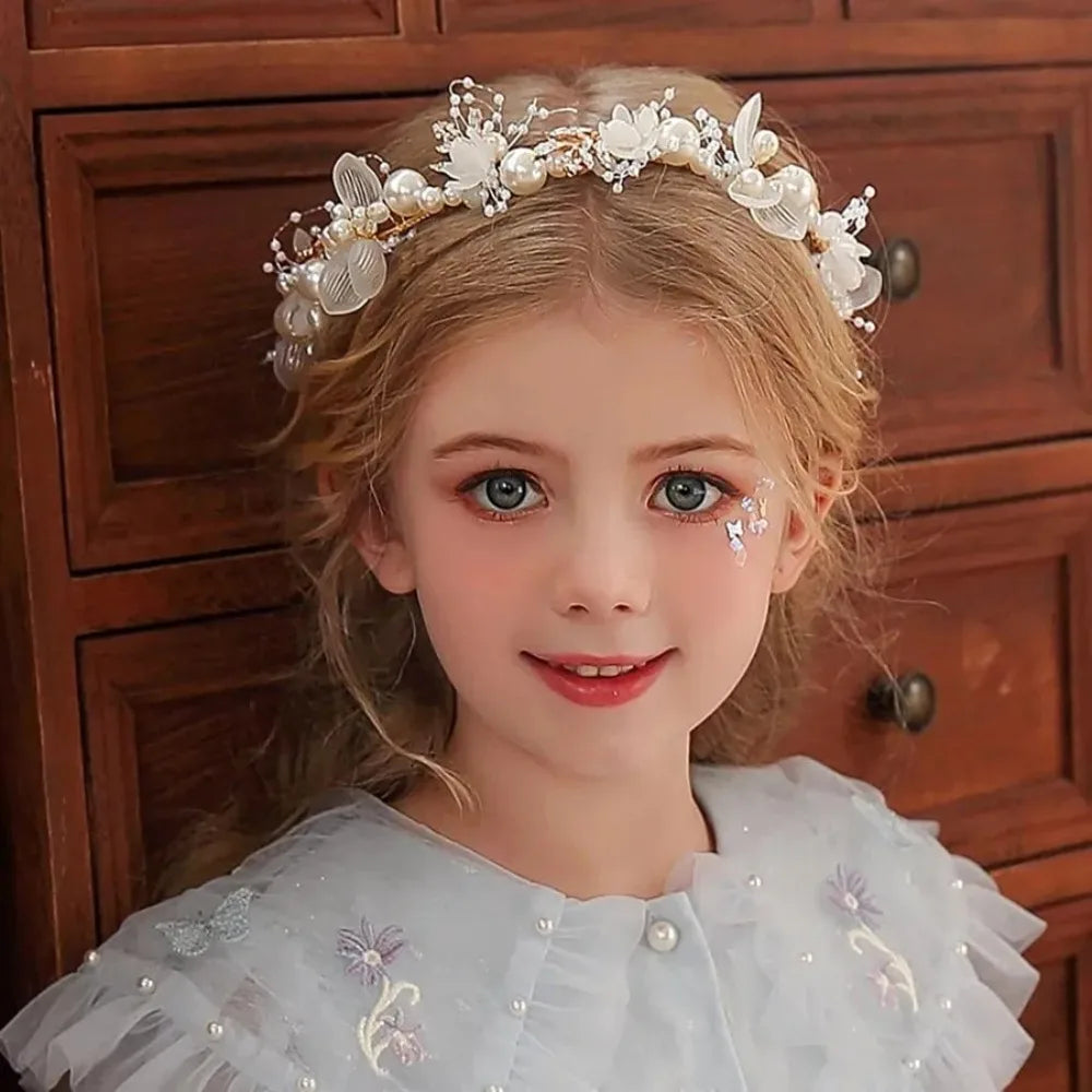 A young girl wearing an elegant floral wreath headband and a white dress, smiling at the camera.