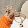 Una mano sostiene gafas cuadradas transparentes con detalles dorados sobre un fondo blanco.