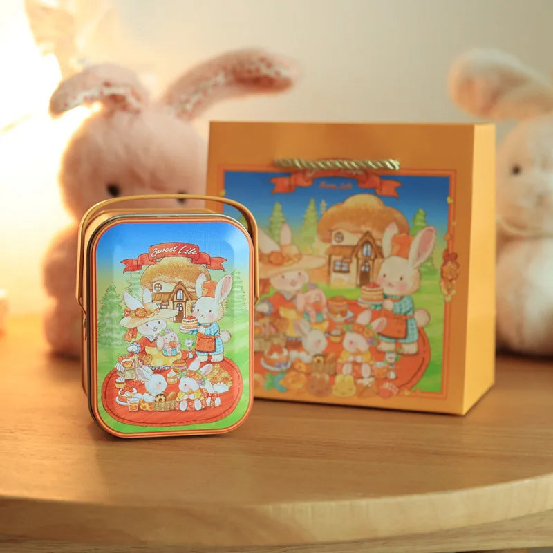 An orange tinplate candy box featuring a scene of rabbits and a house, with a carrying loop, next to a fluffy pink rabbit.