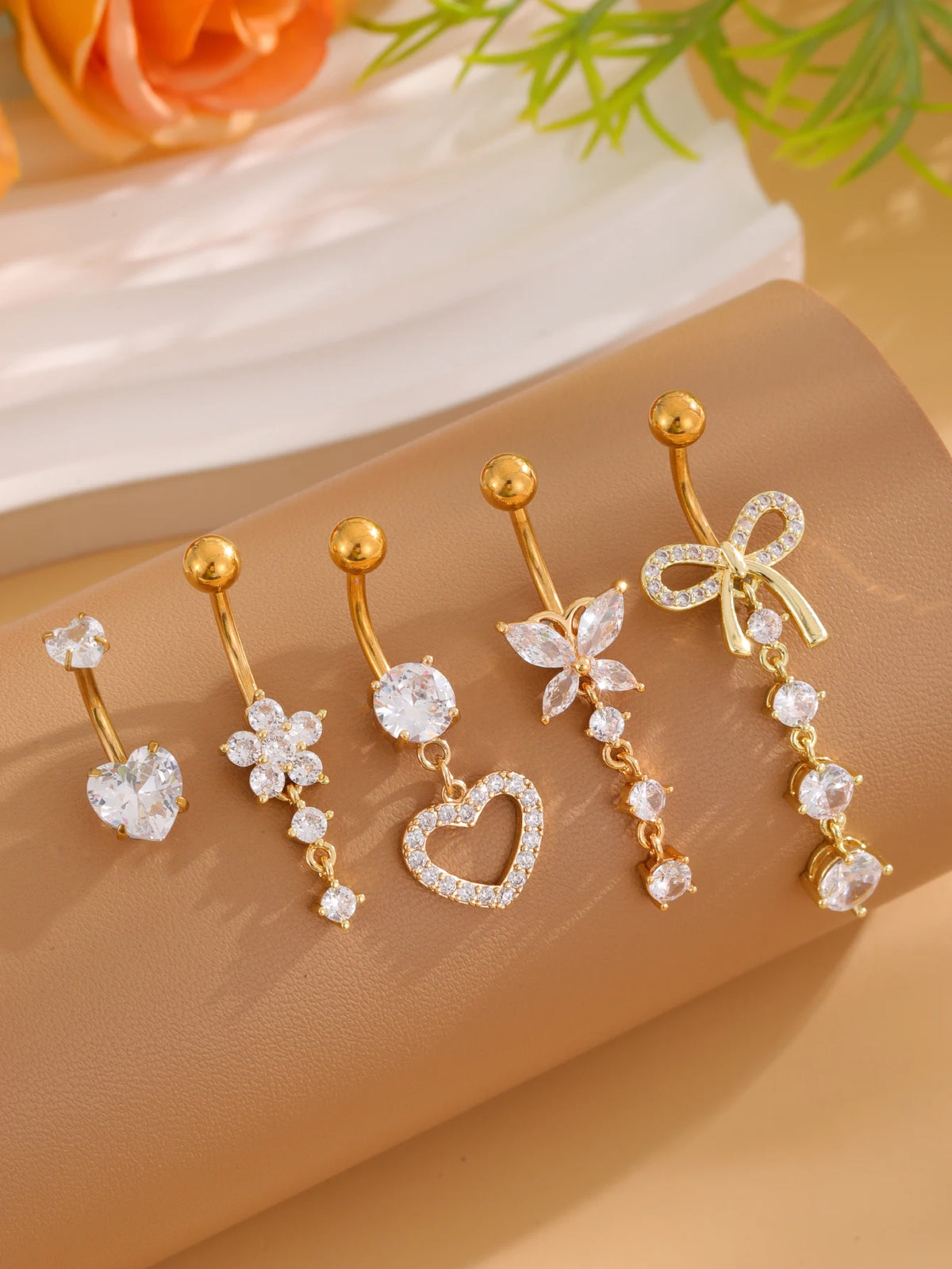 A set of five gold stainless steel dangled belly button rings with various charms and designs displayed on a beige surface.