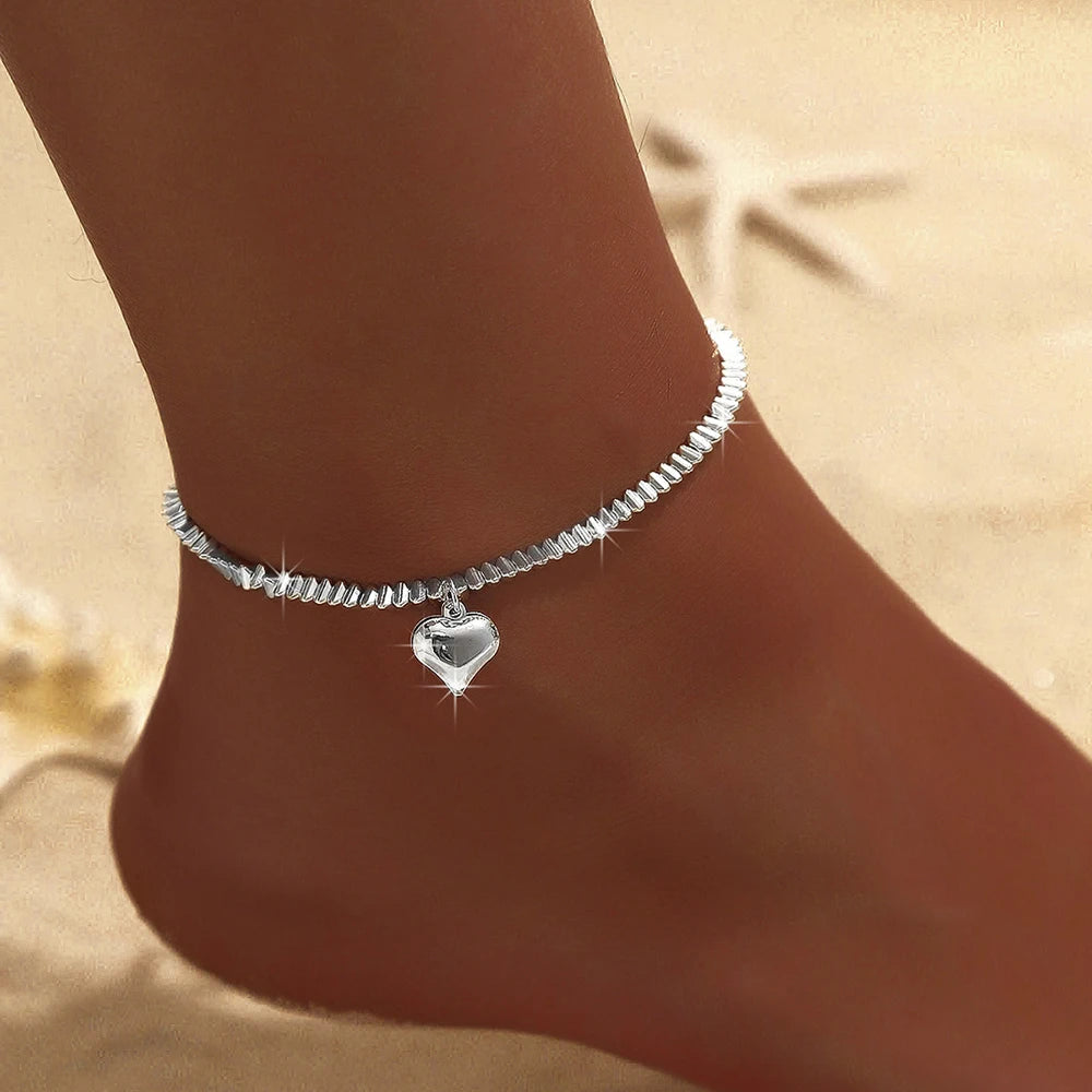 A close-up of a sparkling zircon heart cross anklet worn on a person's ankle.