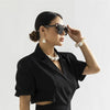 Une femme portant un blazer noir et des lunettes est posant, mettant en valeur un élégant collier imitation perles.